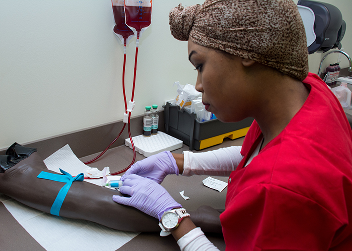 A female phlebotomist sticks a prosthetic training arm in phlebotomy class.